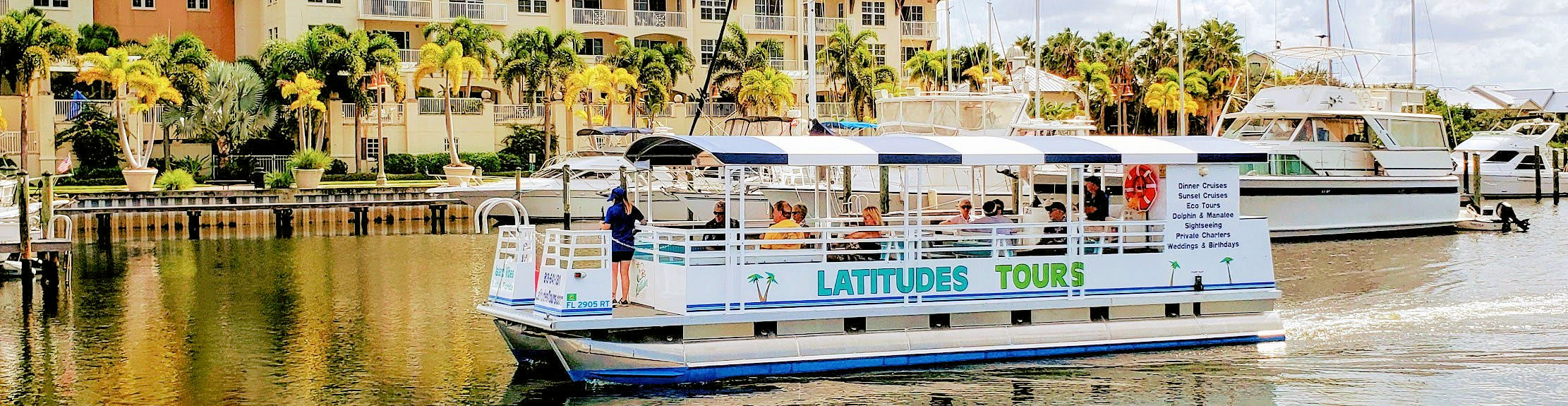 A tour boat named 'Latitudes Tours' with passengers aboard, navigating a calm waterway lined with palm trees and residential buildings in the background.